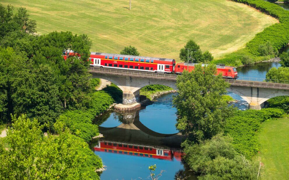 Regionalbahn fährt über eine Brücke in grüner Natur. Im Fluss unter der Brücke spiegeln sich Brücke und Bahn